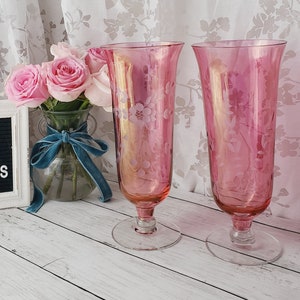 May include: Two pink glass vases with etched floral designs. The vases have a clear glass base and are sitting on a white wooden surface. A sign with the text "Hello Gorgeous" is in the background.