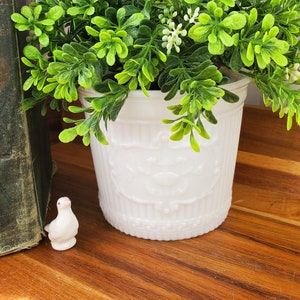 May include: A white milk glass planter with a raised design, filled with a green leafy plant. The planter is sitting on a wooden surface with a stack of old books and a white ceramic bird figurine.