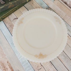 May include: A white milk glass dinner plate with a gold floral design. The plate is on a light blue and white wooden surface.