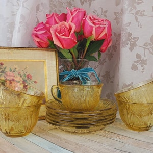 May include: A set of six amber glass teacups and saucers with a textured design. The cups and saucers are arranged on a wooden surface with a bouquet of pink roses in the background.
