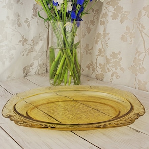 May include: A yellow glass serving platter with a decorative pattern. The platter is on a white wooden surface. A bouquet of white and blue flowers is in a glass vase behind the platter.