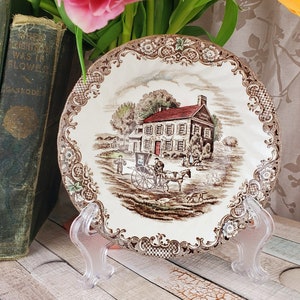 May include: A brown and white ceramic plate with a vintage scene of a house with a red roof and a horse-drawn carriage. The plate is on a clear plastic stand. The plate has a gold trim and a floral design.