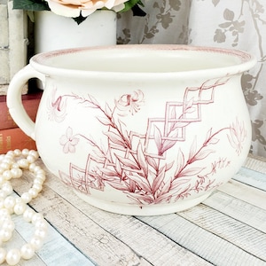 May include: A white ceramic chamber pot with a red floral design. The pot has two handles and a rounded shape.