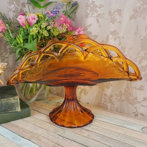 May include: Amber glass pedestal bowl with a woven design. The bowl is on a wooden surface with a seashell and books.