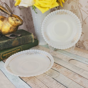 May include: Two white milk glass plates with gold trim and a scalloped edge. The plates are on a wooden surface with a gold rabbit figurine and a stack of books in the background.
