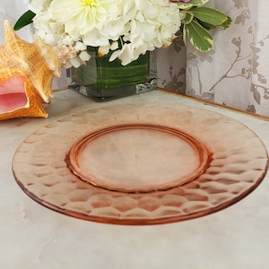 Puede incluir: Un plato de vidrio rosa con un patrón texturizado en forma de panal. El plato es redondo y tiene un borde ligeramente elevado. Una concha marina y un arreglo floral con flores blancas, rosas y verdes están en el fondo.