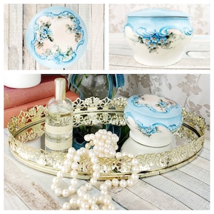 May include: A gold mirrored tray with a decorative lace-like edge, featuring a pearl necklace, a bottle of perfume, and two blue and white porcelain trinket boxes with floral designs.