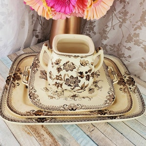 May include: Three vintage ceramic serving trays with a floral pattern in brown on a cream background. The trays are stacked on top of each other and have a decorative border. A cream ceramic pitcher with a floral pattern in brown is sitting on top of the trays.