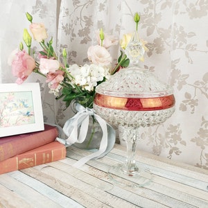 May include: A clear glass candy dish with a red rim and a diamond pattern. The dish is on a wooden table with a bouquet of pink and white flowers in a vase behind it. A white picture frame with a floral print and the text "Here we pause to think of those we love" is on the table in front of the dish.