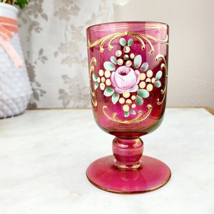 May include: A vintage stemmed glass with a pink and gold design. The glass has a floral pattern with a pink rose and green leaves. The glass is decorated with gold trim and dots.