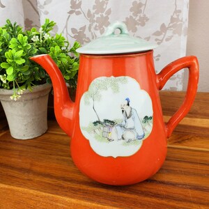 May include: A red ceramic teapot with a white lid and a painted scene of a person sitting under a tree. The teapot has a gold trim around the lid and the painted scene.