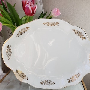 May include: White oval serving platter with a scalloped edge and gold floral accents. The vintage dish has a glossy finish and is decorated with clusters of gold flowers around the rim. A decorative piece for serving food.