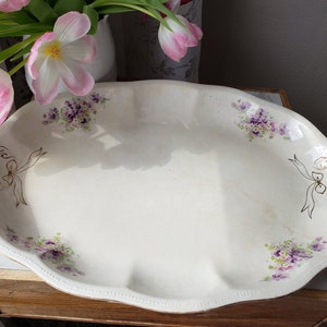 May include: A white oval serving dish with a scalloped edge. The dish is decorated with a floral pattern of purple and green flowers and gold ribbon bows.