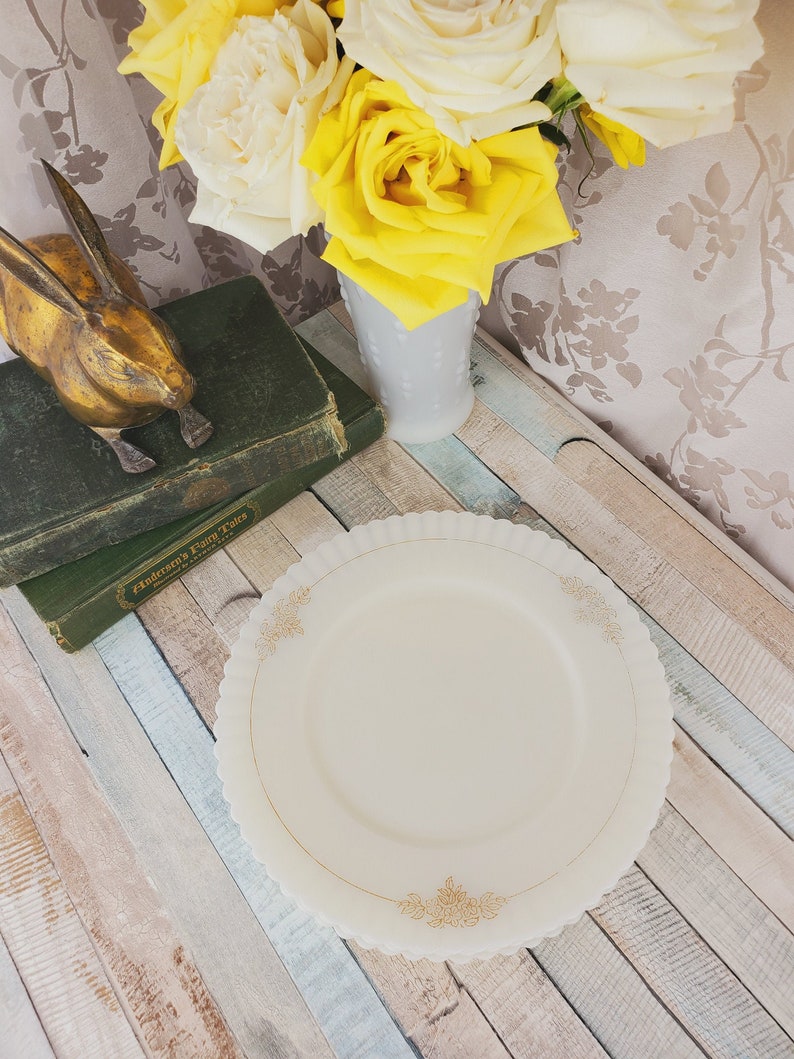 May include: A white milk glass dinner plate with a gold floral design. The plate is on a light blue and white wooden surface.