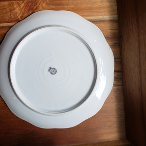 K&ouml;nnte beinhalten: Wei&szlig;e Porzellanplatte mit gewelltem Rand. Die Unterseite der Platte hat ein schwarz-wei&szlig;es Logo mit einer Krone.