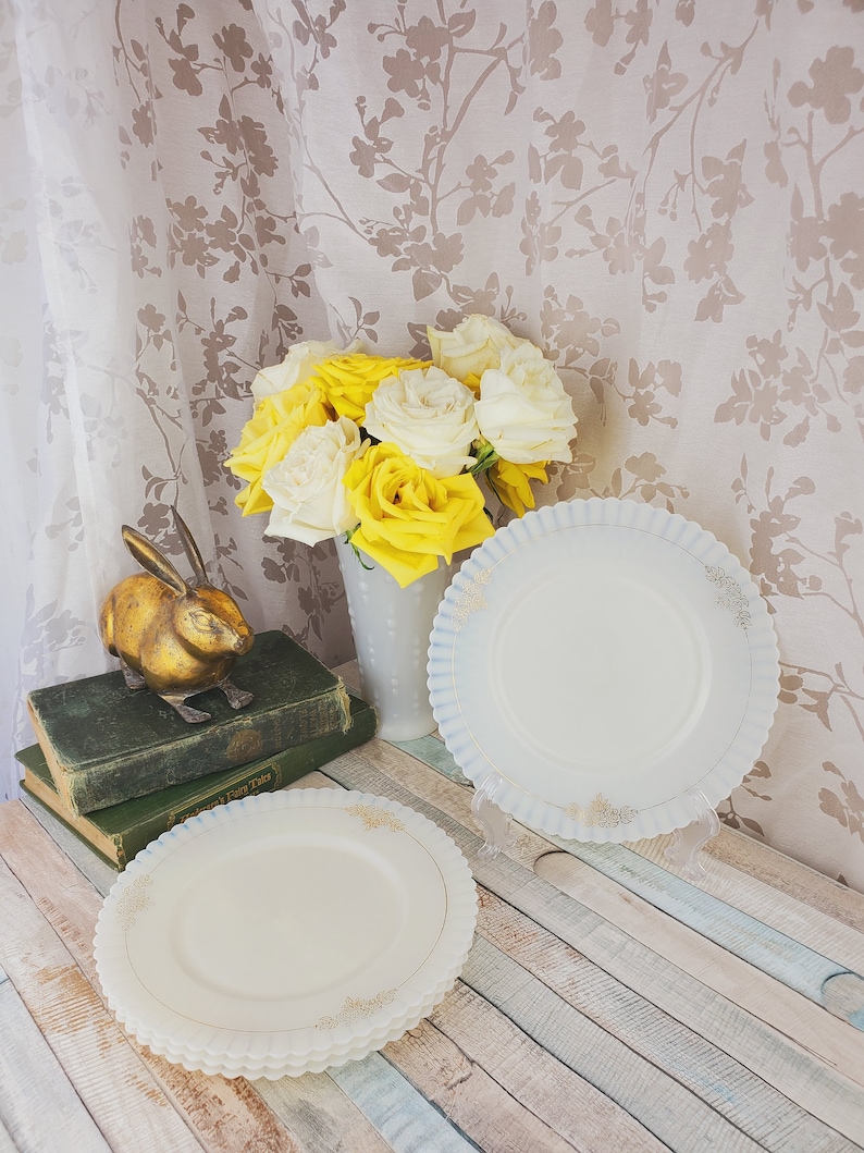 May include: Two white milk glass plates with a scalloped edge and gold trim. The plates are stacked on a wooden surface. A bouquet of yellow and white roses is in the background.