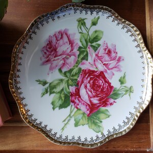 K&ouml;nnte beinhalten: Ein wei&szlig;er Porzellanteller mit goldfarbenem Rand und einem Blumendesign mit rosa und roten Rosen mit gr&uuml;nen Bl&auml;ttern.