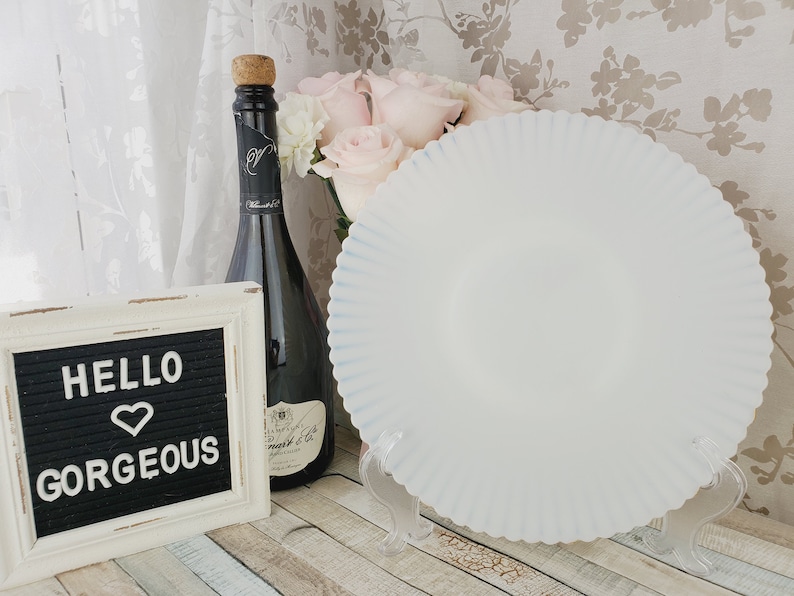 K&ouml;nnte beinhalten: Ein wei&szlig;er, geriffelter Keramikteller mit blauem Rand steht auf einem durchsichtigen St&auml;nder. Der Teller befindet sich auf einer Holzoberfl&auml;che mit einem schwarzen Buchstabenbrett, auf dem "Hello Gorgeous" steht, und einer Flasche Champagner.