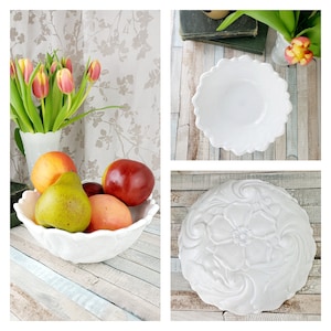 May include: Three white ceramic bowls with different designs. The bowl on the left has a scalloped edge and is filled with fruit. The bowl in the middle has a scalloped edge and is empty. The bowl on the right has a floral design and is empty.