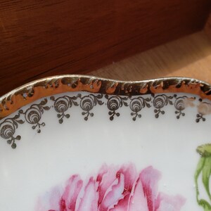 K&ouml;nnte beinhalten: Ein wei&szlig;er Porzellanteller mit goldfarbenem Rand und einem schwarzen Blumendesign. Die Mitte des Tellers zeigt eine rosa Rose.