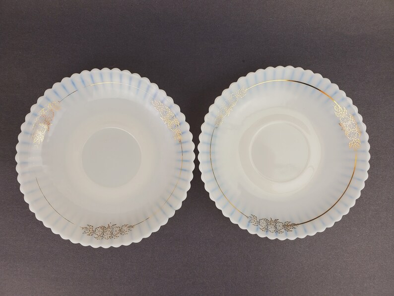 May include: Two white, scalloped-edge saucers with a blue and gold floral design. The saucers are made of opaque glass and have a raised center.