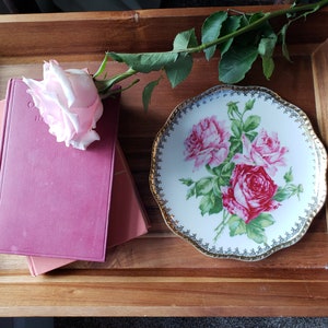 K&ouml;nnte beinhalten: Ein Holztablett mit einer rosa Rose, einem Stapel von zwei B&uuml;chern und einem wei&szlig;en Porzellanteller mit einem Blumendesign. Der Teller hat einen goldenen Rand und ein goldenes Blumenmuster.