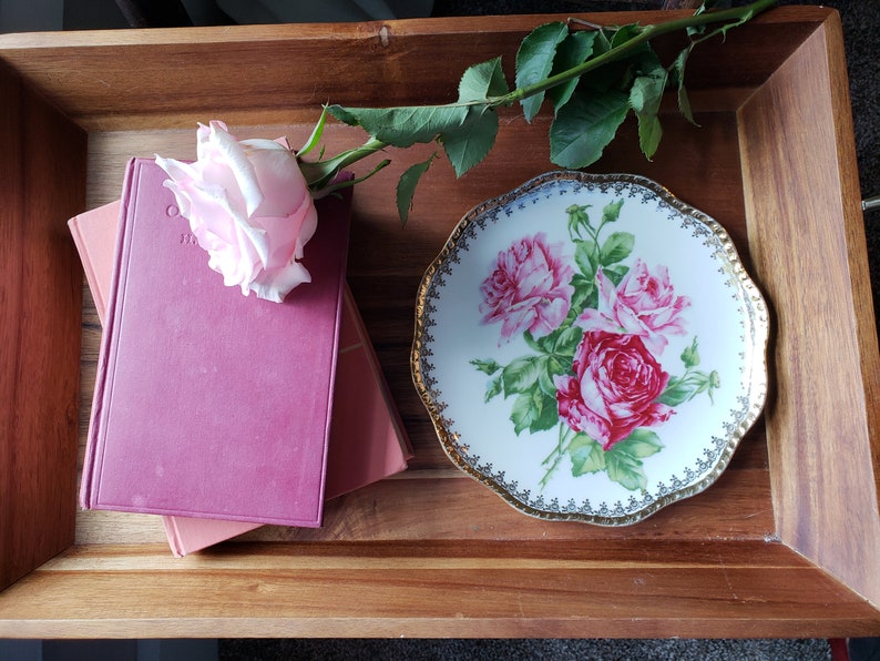 K&ouml;nnte beinhalten: Ein Holztablett mit einer rosa Rose, einem Stapel von zwei B&uuml;chern und einem wei&szlig;en Porzellanteller mit einem Blumendesign. Der Teller hat einen goldenen Rand und ein goldenes Blumenmuster.