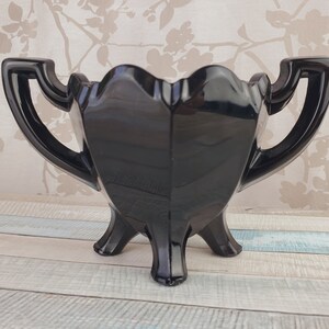 K&ouml;nnte beinhalten: Eine schwarze Glasvase mit zwei Griffen und drei Beinen. Die Vase hat eine herzf&ouml;rmige Form und eine gl&auml;nzende Oberfl&auml;che.