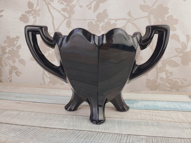 K&ouml;nnte beinhalten: Eine schwarze Glasvase mit zwei Griffen und drei Beinen. Die Vase hat eine herzf&ouml;rmige Form und eine gl&auml;nzende Oberfl&auml;che.