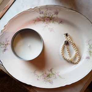 May include: A white candle in a silver tin and a pearl necklace on a white oval platter with a floral pattern.