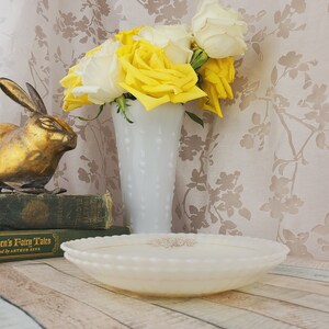 May include: A gold rabbit figurine sits on a stack of books with the title "Anderson's Fairy Tales" visible.  A white milk glass vase with a textured surface holds a bouquet of yellow and white roses.