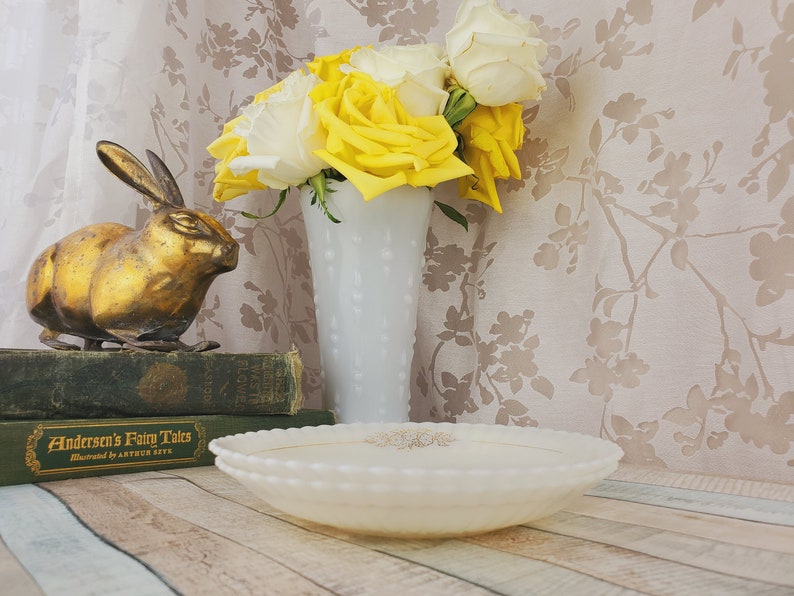 May include: A gold rabbit figurine sits on a stack of books with the title "Anderson's Fairy Tales" visible.  A white milk glass vase with a textured surface holds a bouquet of yellow and white roses.