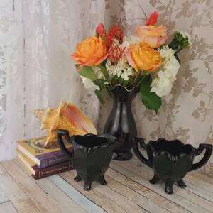 K&ouml;nnte beinhalten: Ein Set aus zwei schwarzen Glas-Sahnegie&szlig;ern und Zuckerbeh&auml;ltern mit geriffelten R&auml;ndern und drei Beinen. Die Schalen stehen auf einer Holzoberfl&auml;che mit einer Muschel und B&uuml;chern.