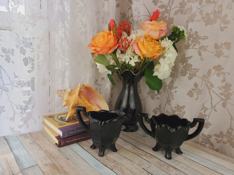 K&ouml;nnte beinhalten: Ein Set aus zwei schwarzen Glas-Sahnegie&szlig;ern und Zuckerbeh&auml;ltern mit geriffelten R&auml;ndern und drei Beinen. Die Schalen stehen auf einer Holzoberfl&auml;che mit einer Muschel und B&uuml;chern.