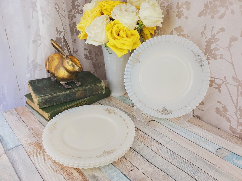 May include: Two white vintage milk glass plates with a blue and gold trim. The plates are stacked on a wooden surface.