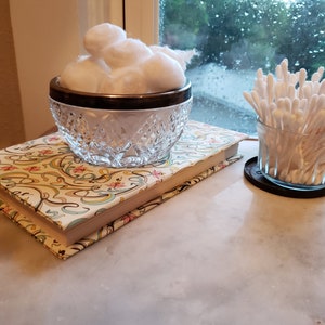 May include: A clear, cut-glass bowl with a silver rim filled with fluffy cotton balls sits atop a decorative book with a floral pattern. A small glass jar of cotton swabs rests on a coaster nearby.