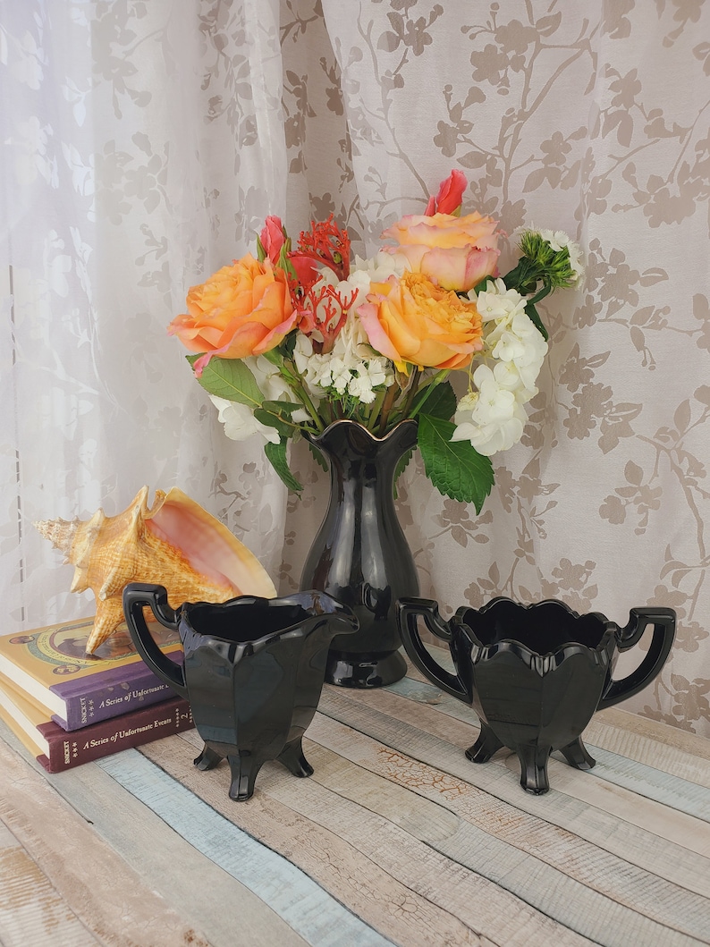 K&ouml;nnte beinhalten: Ein Set aus zwei schwarzen Glas-Sahnegie&szlig;ern und Zuckerbeh&auml;ltern mit gewelltem Rand und drei Beinen. Die Schalen stehen auf einer Holzoberfl&auml;che mit einer Muschel und B&uuml;chern im Hintergrund. Ein Strau&szlig; orangefarbener und wei&szlig;er Blumen in einer schwarzen Vase befindet sich hinter den Schalen.