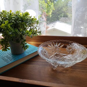 Könnte beinhalten: Eine klare Glasschale auf einem Sockel mit einem Sternmuster steht auf einem Holztablett mit einer kleinen Topfpflanze und einem blauen Buch mit dem Titel "Jane Austen" darauf.