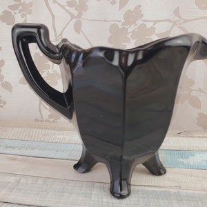 K&ouml;nnte beinhalten: Eine schwarze Glas-Sahnegie&szlig;kanne mit einem dreibeinigen Sockel und einem Griff. Die Kanne ist aus gl&auml;nzendem schwarzem Glas und hat eine glatte, abgerundete Form.