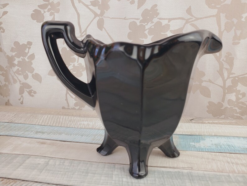 K&ouml;nnte beinhalten: Eine schwarze Glas-Sahnegie&szlig;kanne mit einem dreibeinigen Sockel und einem Griff. Die Kanne ist aus gl&auml;nzendem schwarzem Glas und hat eine glatte, abgerundete Form.