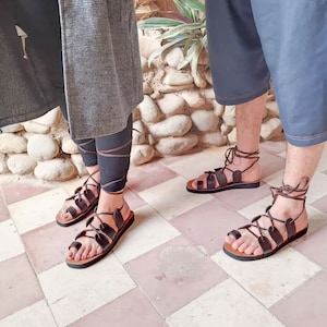 May include: Brown leather sandals with multiple straps and a toe loop. The sandals have a dark brown sole and are laced up the ankle with brown laces. The sandals are worn by two people.