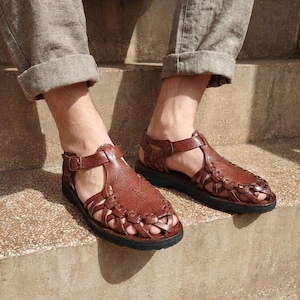 Leather Fisherman Sandals: Women's Beige Gladiator Closed Toe Shoes
handcrafted, brown leather huarache-style sandals. The sandals feature a smooth, polished leather shield over the top of the foot with decorative embossed lines, transitioning into a woven latticework design around the toes. They have adjustable ankle straps with brass-tone buckles and thick, black rubber soles