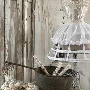 May include: A vintage metal bowl filled with rolled sheet music, sitting on a white lace tablecloth. A white crinoline skirt and a white lace bodice are in the background.