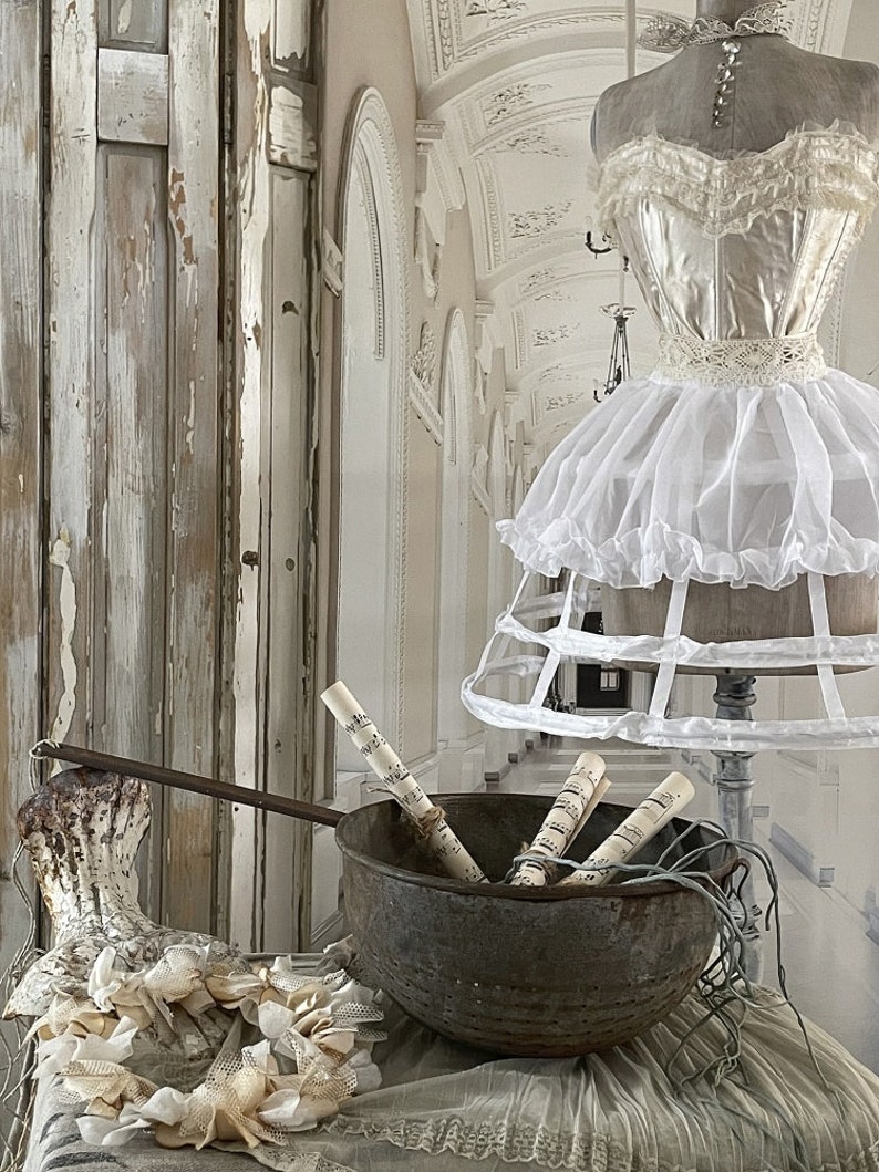 May include: A vintage metal bowl filled with rolled sheet music, sitting on a white lace tablecloth. A white crinoline skirt and a white lace bodice are in the background.