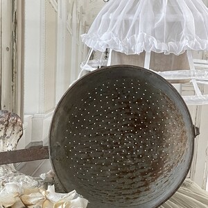May include: A large, round, metal colander with a handle. The colander is made of a dark metal with a speckled finish. It is sitting on a white surface.