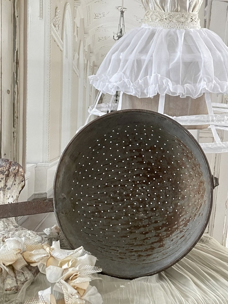 May include: A large, round, metal colander with a handle. The colander is made of a dark metal with a speckled finish. It is sitting on a white surface.