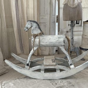 Könnte beinhalten: Ein weißes Schaukelpferd aus Holz im Shabby-Chic-Stil mit grauer Mähne und Schweif. Das Pferd hat einen Sattel und ist mit Bindfaden gebunden. Die geschwungenen Kufen sind weiß lackiert. Ein dekorativer Artikel.