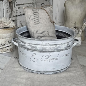 May include: A white, distressed metal bucket with handles, inscribed with "Eau de Lavande". Inside, a linen sack is tied with twine. Other vintage items are in the background, including a hat box and a dress form.