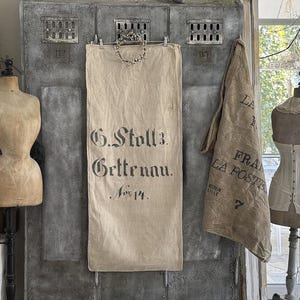 Könnte beinhalten: Ein rechteckiger beigefarbener Jutesack hängt an einem grauen Metallspind und trägt den schwarzen Aufdruck G. Stoll 3. Gettenau. Nov 14. Ein kleiner dekorativer Kranz ist am oberen Rand des Sacks befestigt, der zwischen zwei Schneiderpuppen hängt.