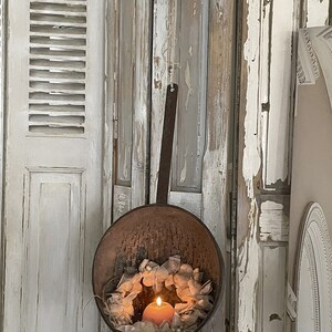 May include: A rustic metal colander hanging from a hook, with a white lace wreath and a lit candle inside. The colander is hanging against a whitewashed wooden wall.